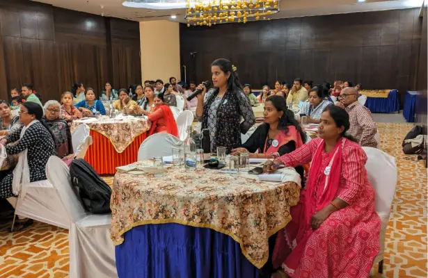 Interactive Discussion at a Professional Conference A woman standing and speaking into a microphone at a conference, with an audience of engaged participants seated at tables in a well-lit conference hall.