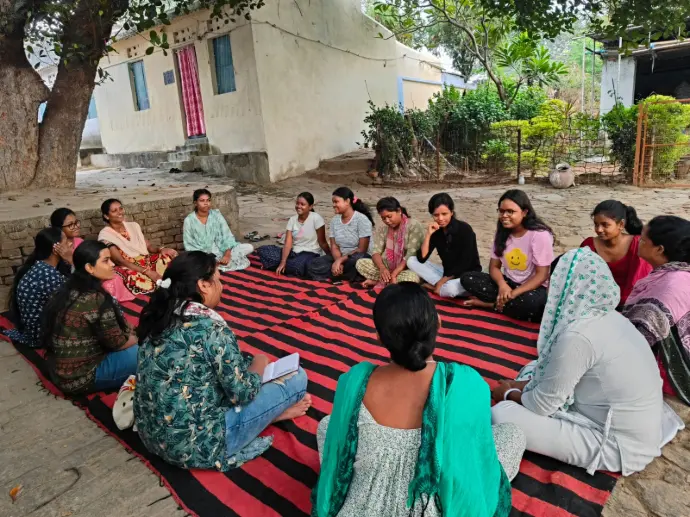 A group of women and adolescent girls sitting in a circle on a striped mat outdoors, engaged in a discussion about healthcare and social rights