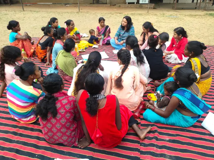Women gathered in a community meeting, engaged in a discussion with a facilitator in an outdoor setting