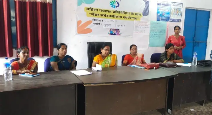Mahila Panchayat representatives participating in a gender sensitization workshop, seated at a conference table with a banner in Hindi in the background.