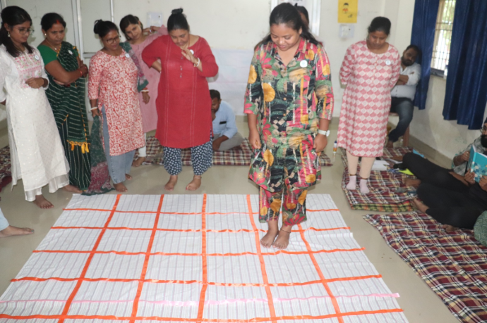 Women Engaged in an Interactive Learning Activity on a Checkered Mat. A group of women gathered around a large checkered mat, with one woman stepping on it as part of an interactive activity in a classroom-like setting. Some participants are observing, while others are seated on the floor with notebooks.